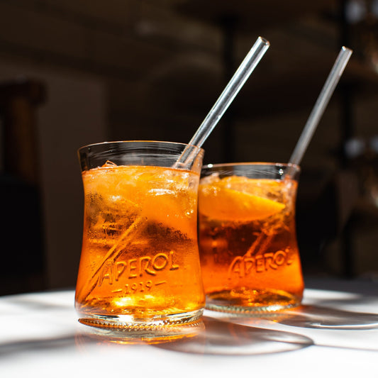 two handmade recycled Aperol glasses sitting on white table with aperol spritz and glass strawes in them