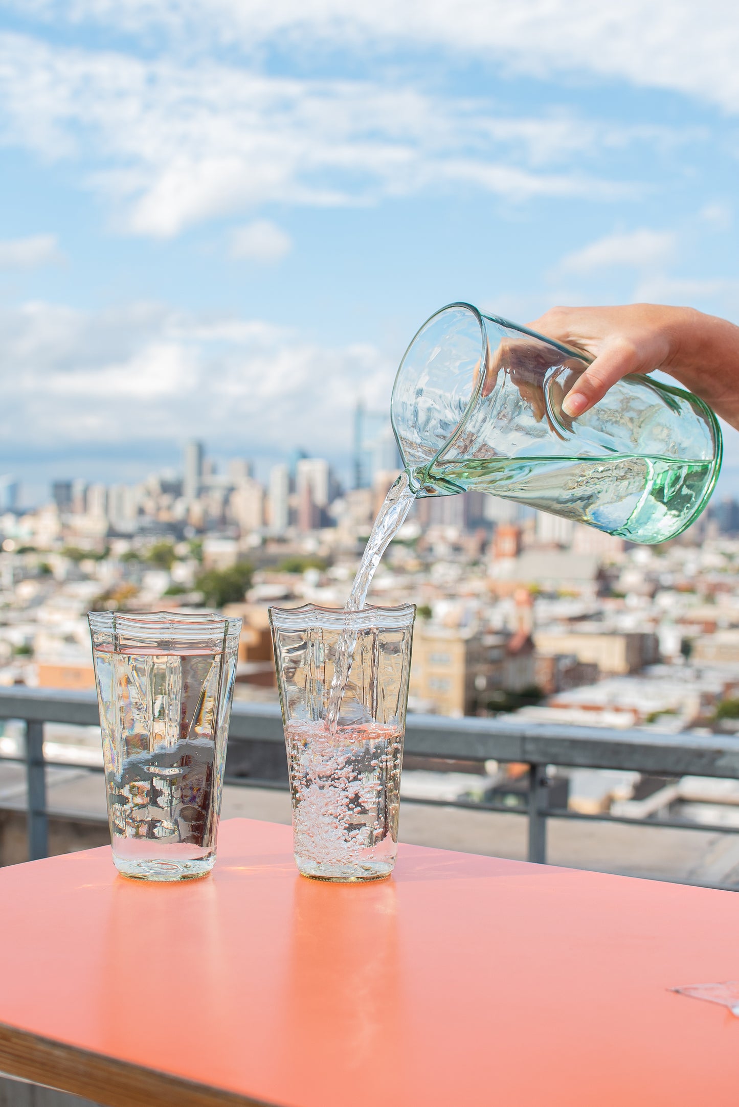 Recycled glass pitcher pouring sparkling water into two recycled water glasses with philadelphia skyline in the background