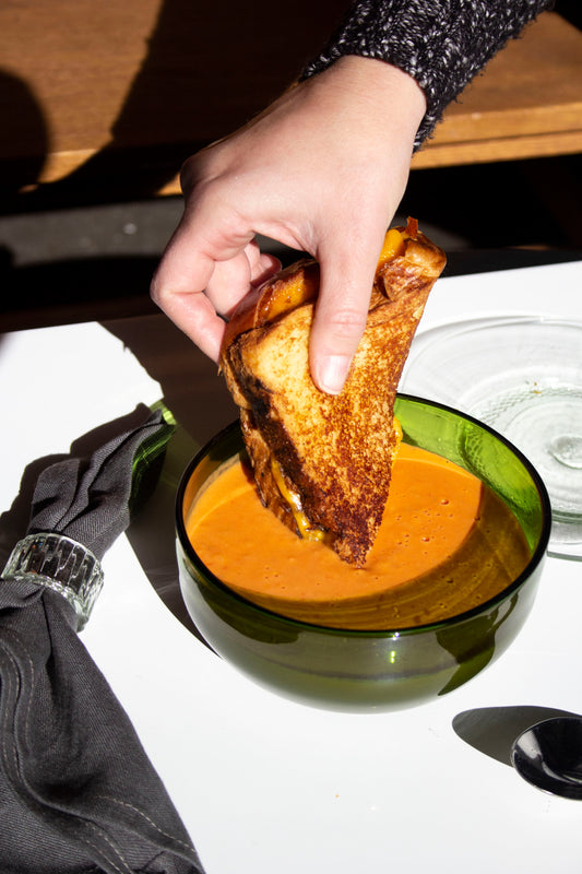 A small recycled glass bowl in the color emerald with a person dipping a grilled cheese into tomato soup.