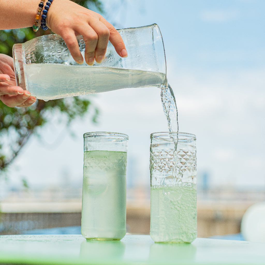 Person pouring a clear liquid from a pitcher into glasses outdoors.