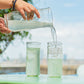 Person pouring a clear liquid from a pitcher into glasses outdoors.