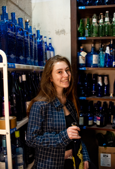 A woman in a blue button up holding a dark blue bottle standing in front of a wall full of blue bottles