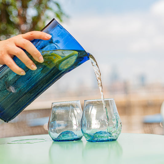 Hand pouring water from a blue glass pitcher into two clear glasses outdoors.