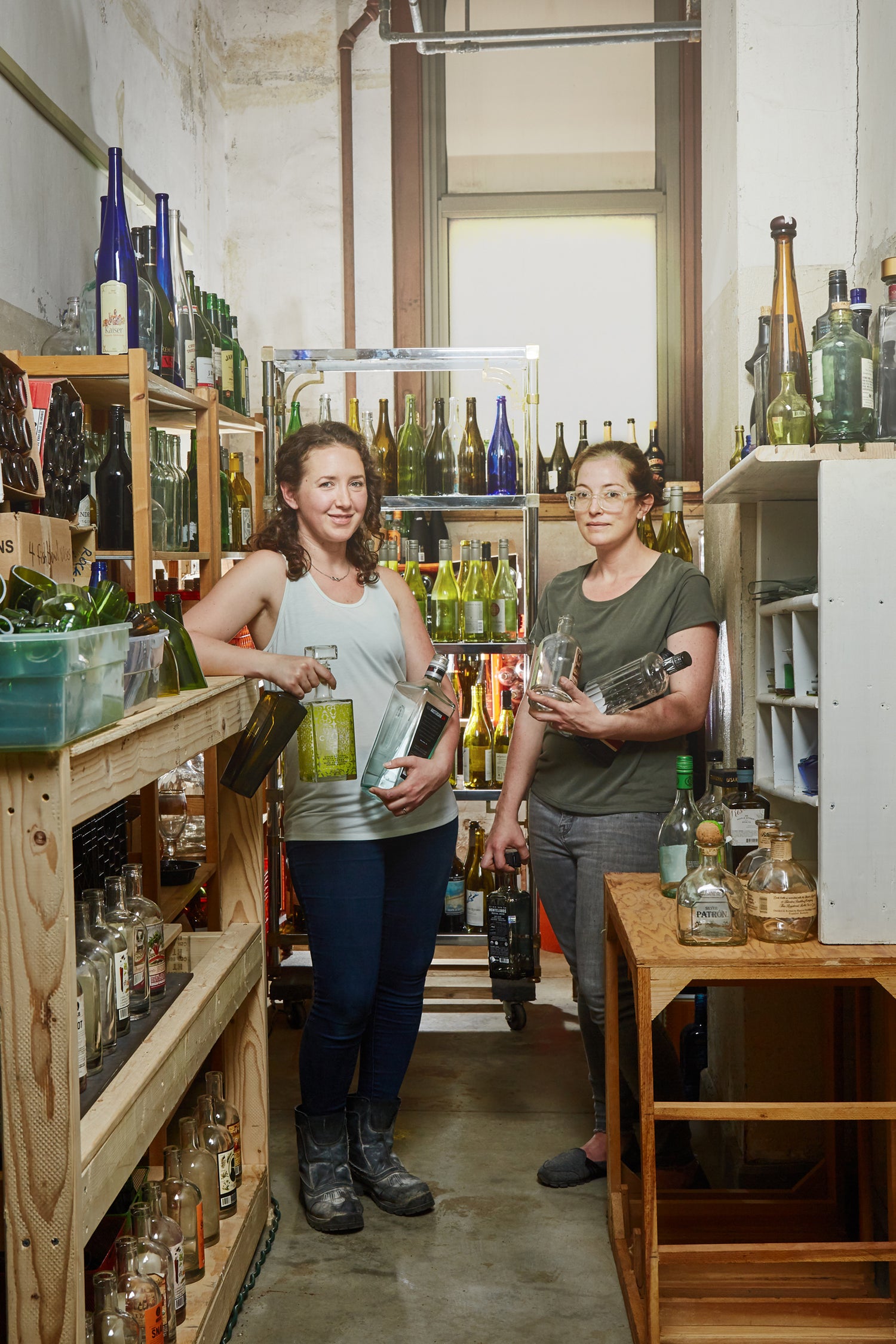 Two women wearing a grey shirt and dark grey shirt holding bottles in a room full of bottles