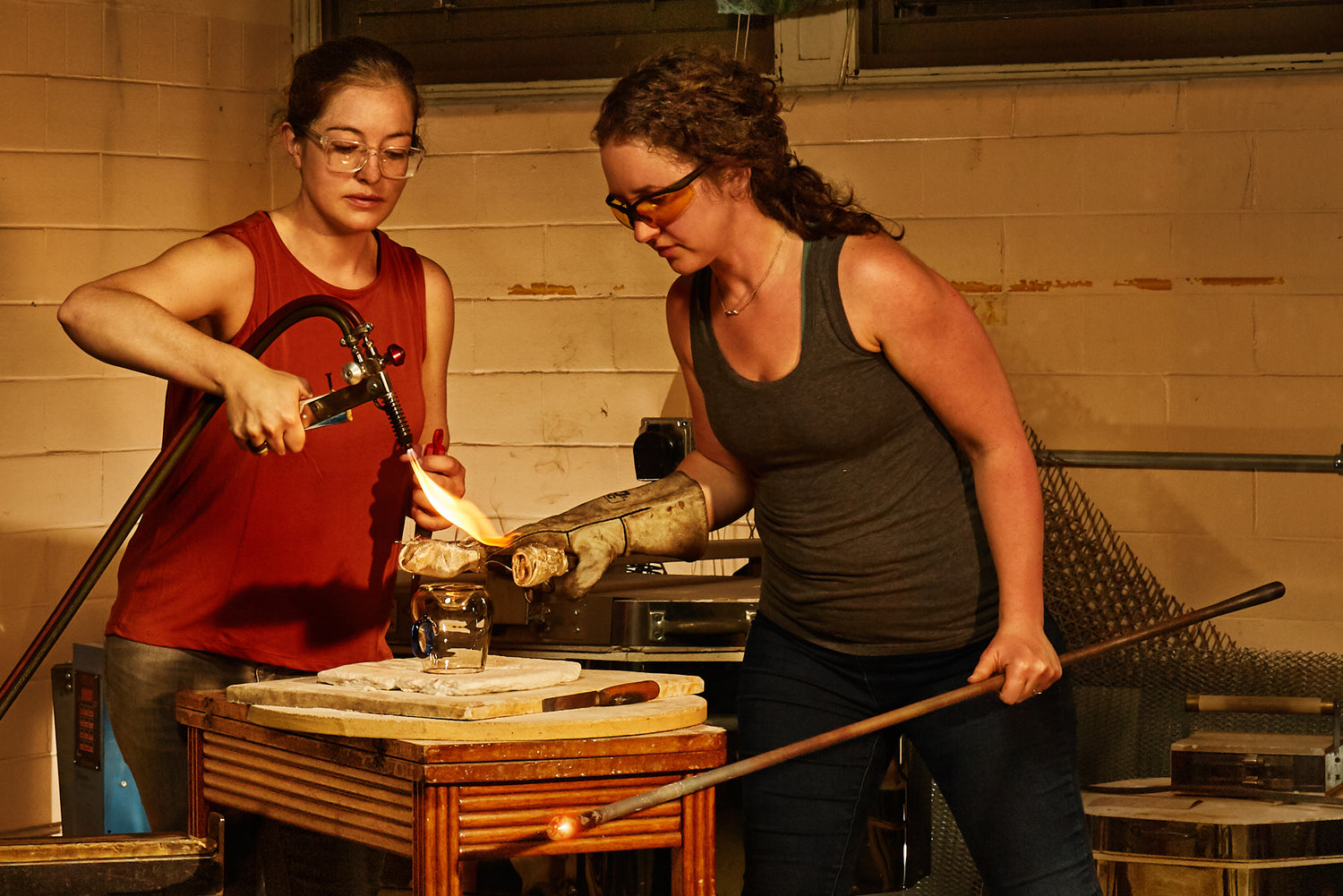 A woman with red shirt using a towch to heat up glass and a woman in grey shirt holding the glass