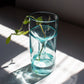 A self-watering planter made from a recycled aqua glass bottle, containing  a young plant. The planter is placed on a windowsill with sunlight shining through