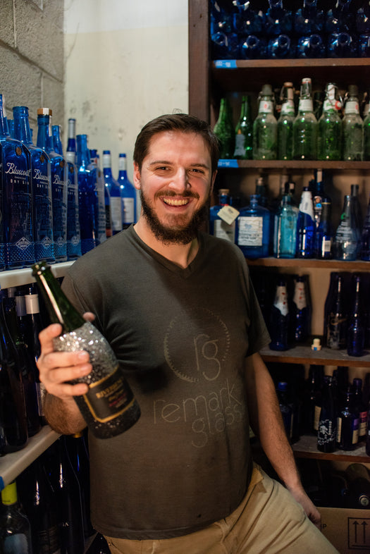 A man in a grey shirt holding a dark bottle standing in front of a wall of blue bottles