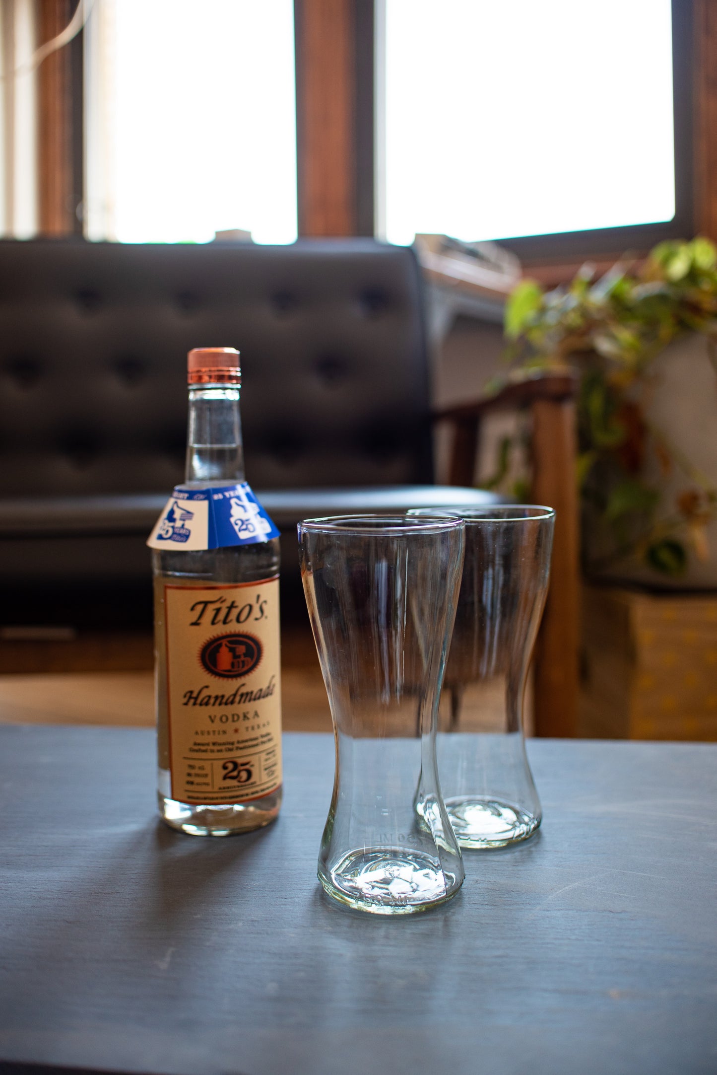 Tito's vodka bottle with two recycled bloody mary glasses sitting on table with a plant and couch
